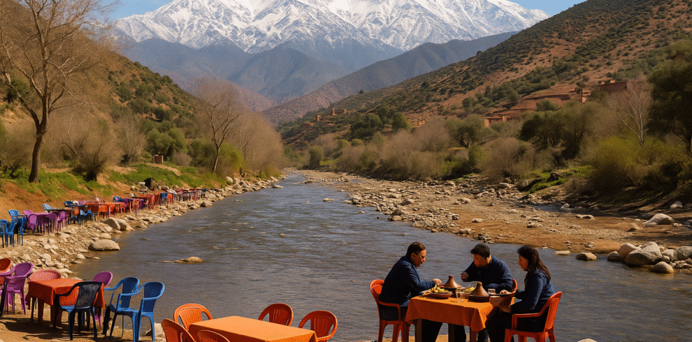 People enjoying a traditional Moroccan tagine meal by the river in Ourika Valley with snow-capped Atlas Mountains in the background