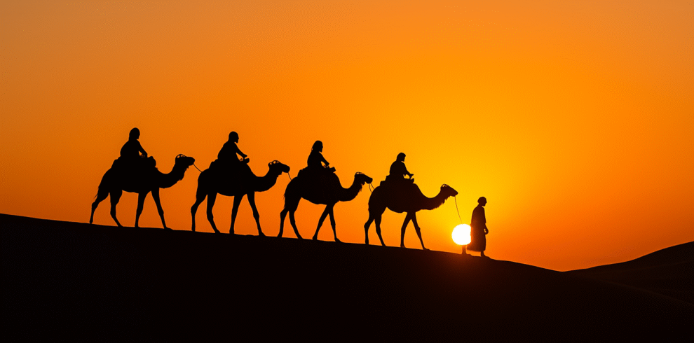 A caravan of camels with riders led by a Berber guide walking across the Merzouga dunes at sunset, captured in a realistic travel photo.morocco small group tours