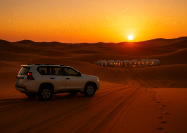 A white 4x4 vehicle parked on the golden dunes of Merzouga with a luxury desert camp glowing in the distance at sunset.morocco small group tours